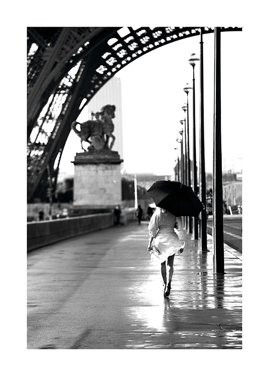  – Fotografía en blanco y negro de una mujer con vestido blanco y paraguas que va caminando al lado de las farolas en la calle que pasa por debajo de la Torre Eiffel
