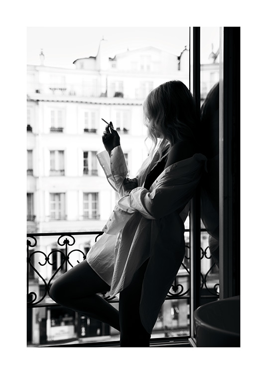  – Fotografía en blanco y negro de una mujer con una camisa extragrande apoyada sobre una ventana y fumando un cigarro