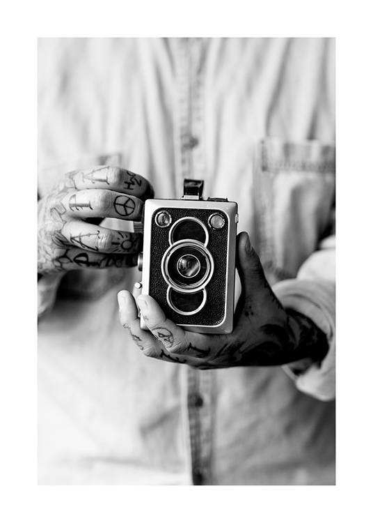  – Fotografía en blanco y negro de un hombre que sostiene una cámara de fotos y tiene las manos tatuadas