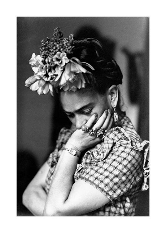  – Fotografía en blanco y negro de Frida Kahlo en una posición pensativa con su rostro apoyado sobre la mano, con camisa a cuadros y flores en la cabeza