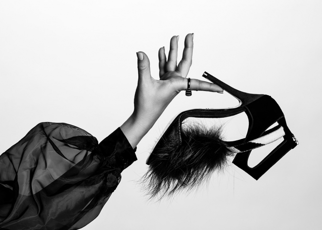  – Fotografía en blanco y negro con la imagen de una brazo femenino que lleva una blusa transparente. La mano sostiene un tacón que tiene un detalle de piel en la punta.