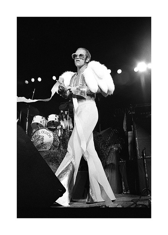  – Fotografía en blanco y negro del cantante Elton John en un escenario. Lleva gafas grandes y está vestido con un mono blanco.
