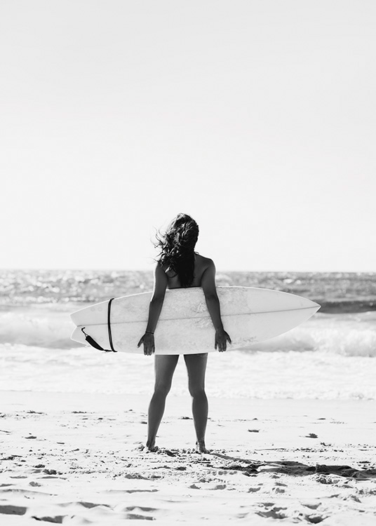  – Fotografía en blanco y negro de una joven de pie en la playa con una tabla de surf mirando hacia el mar.