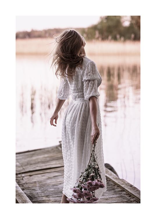  – Fotografía de una mujer con un vestido largo blanco en un muelle pequeño y con un ramillete de flores en la mano.