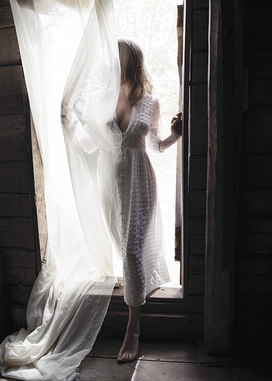  – Fotografía de una mujer con vestido blanco en un umbral con cortina transparente que la cubre al pasarlo.