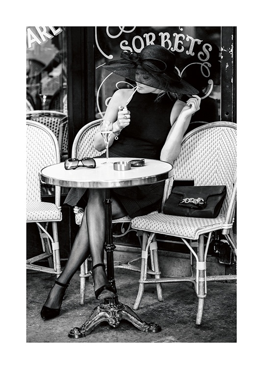 – Fotografía en blanco y negro de una mujer con un sombrero grande fumando un cigarrillo en un café al aire libre.